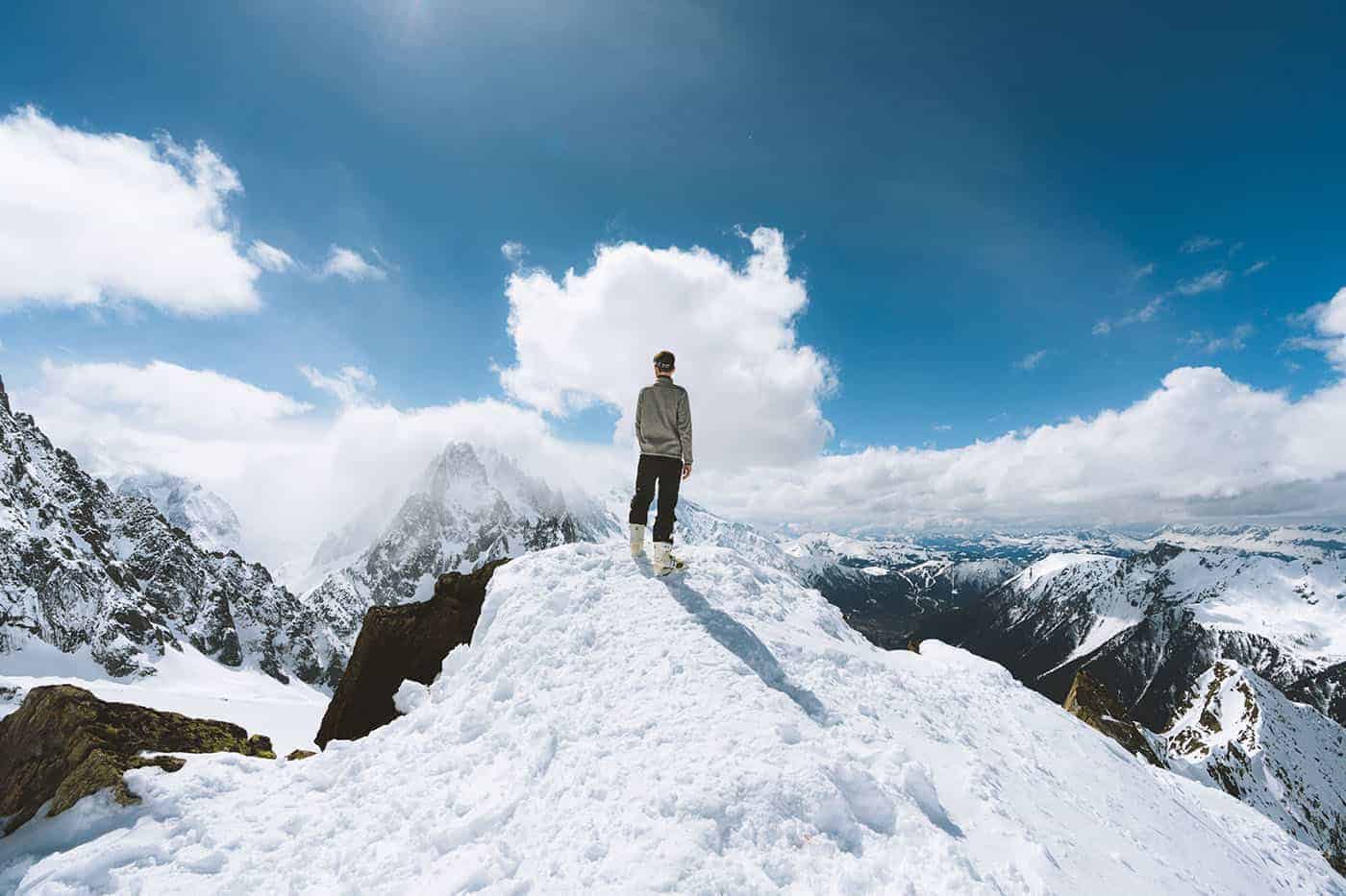 Bergsteiger steht auf verschneiter Gipfelspitze in Alpenkette und blickt über Berglandschaft
